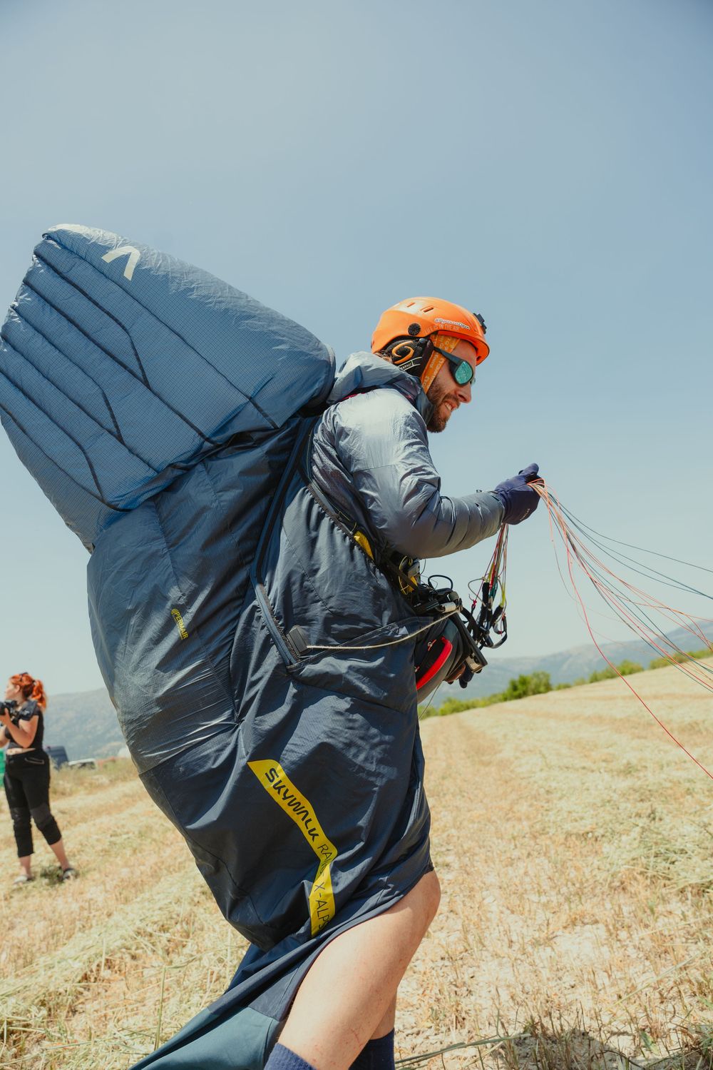 Paraglider in grey flight suit and orange helmet preparing equipment on a dry grassy hill.