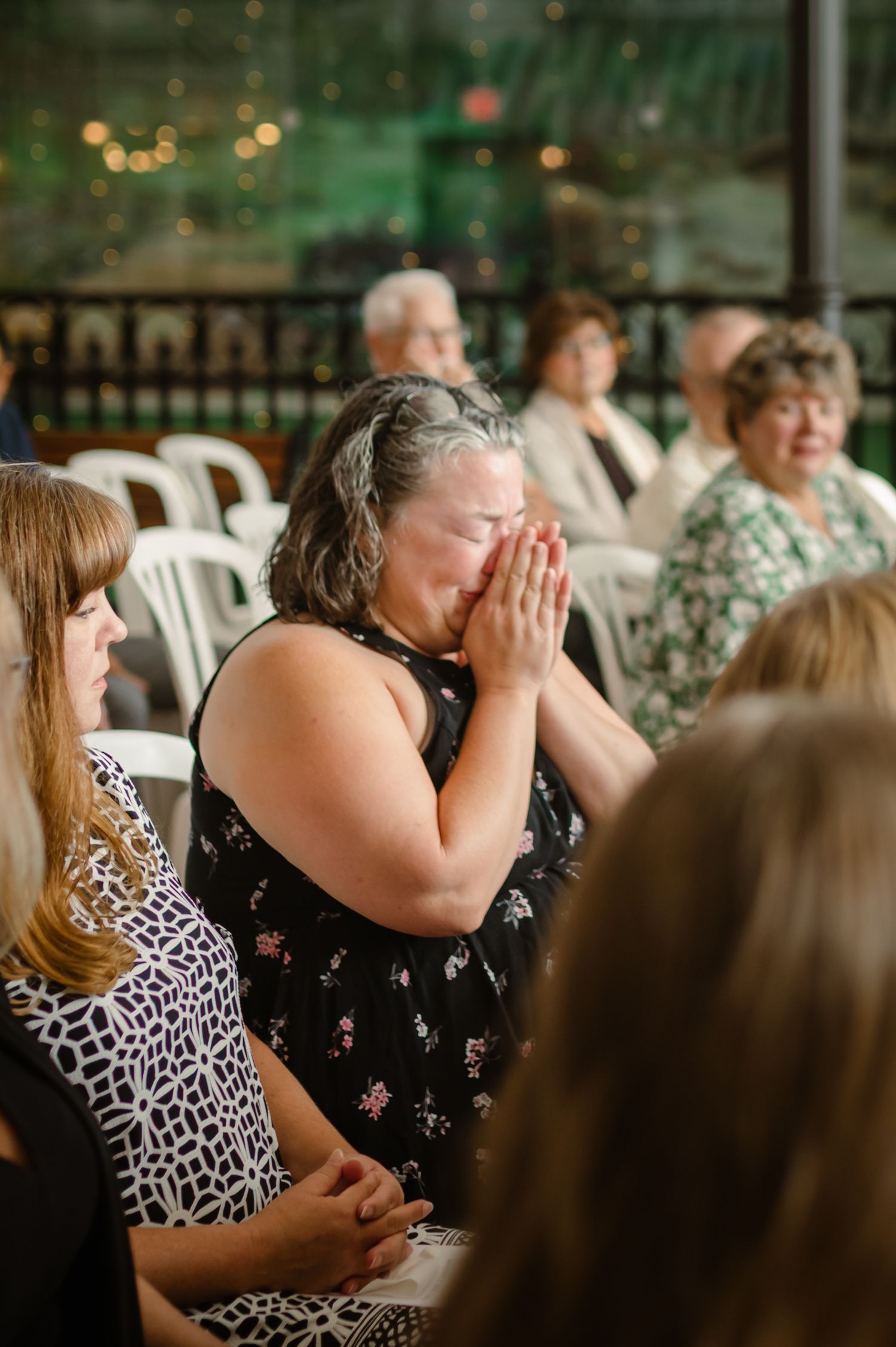 A person in a floral dress covers their face with emotion while sitting among guests at an indoor event.