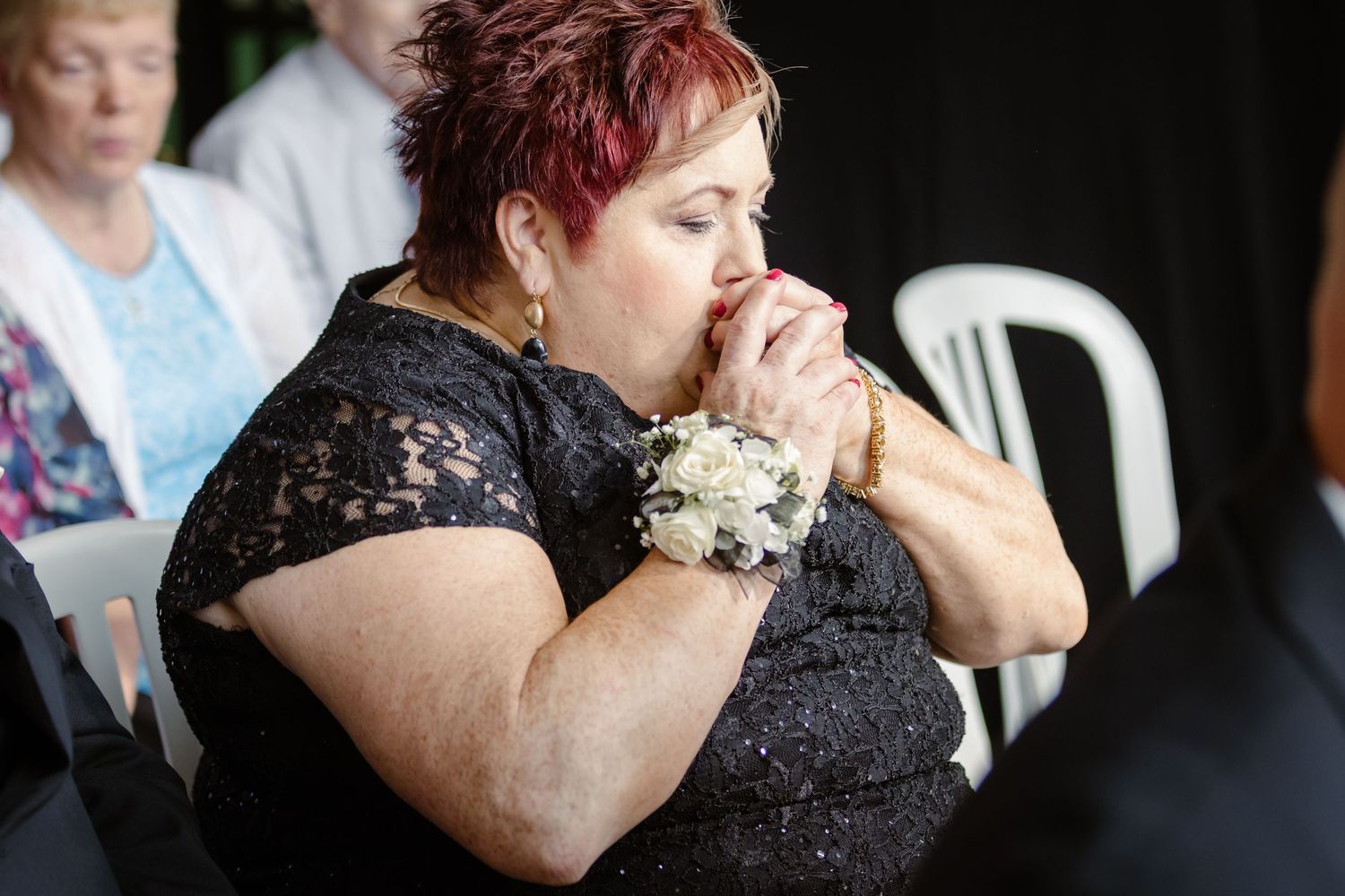 A person in a black lace dress holds a small white bouquet while seated at an event.