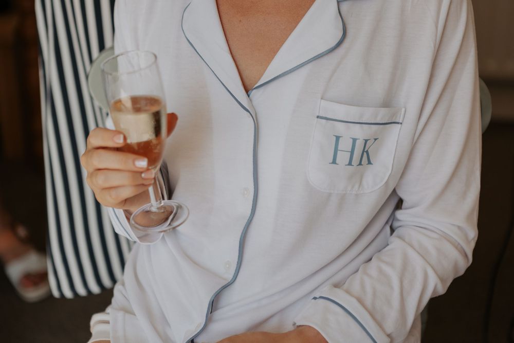 Bride in monogrammed HK white pajamas holding a champagne flute on her wedding morning.