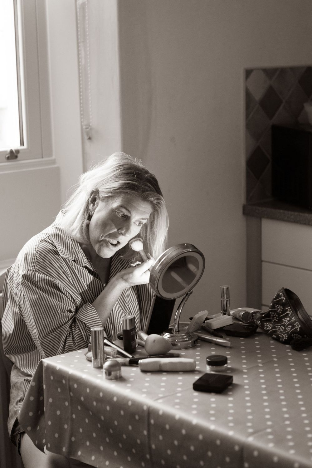Black and white photo of a woman applying makeup at a vanity table with beauty products.