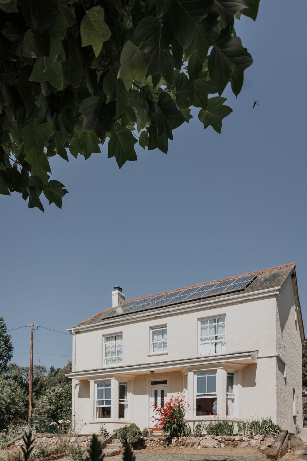 White two-story farmhouse with solar panels on the roof surrounded by trees under a clear blue sky.