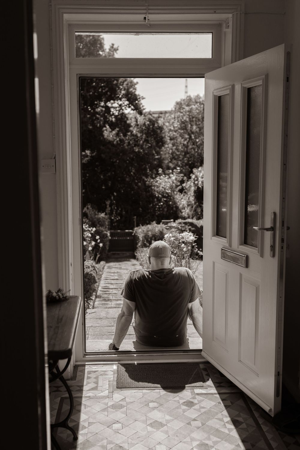 Black and white photo of a man sitting in an open doorway overlooking a lush garden.