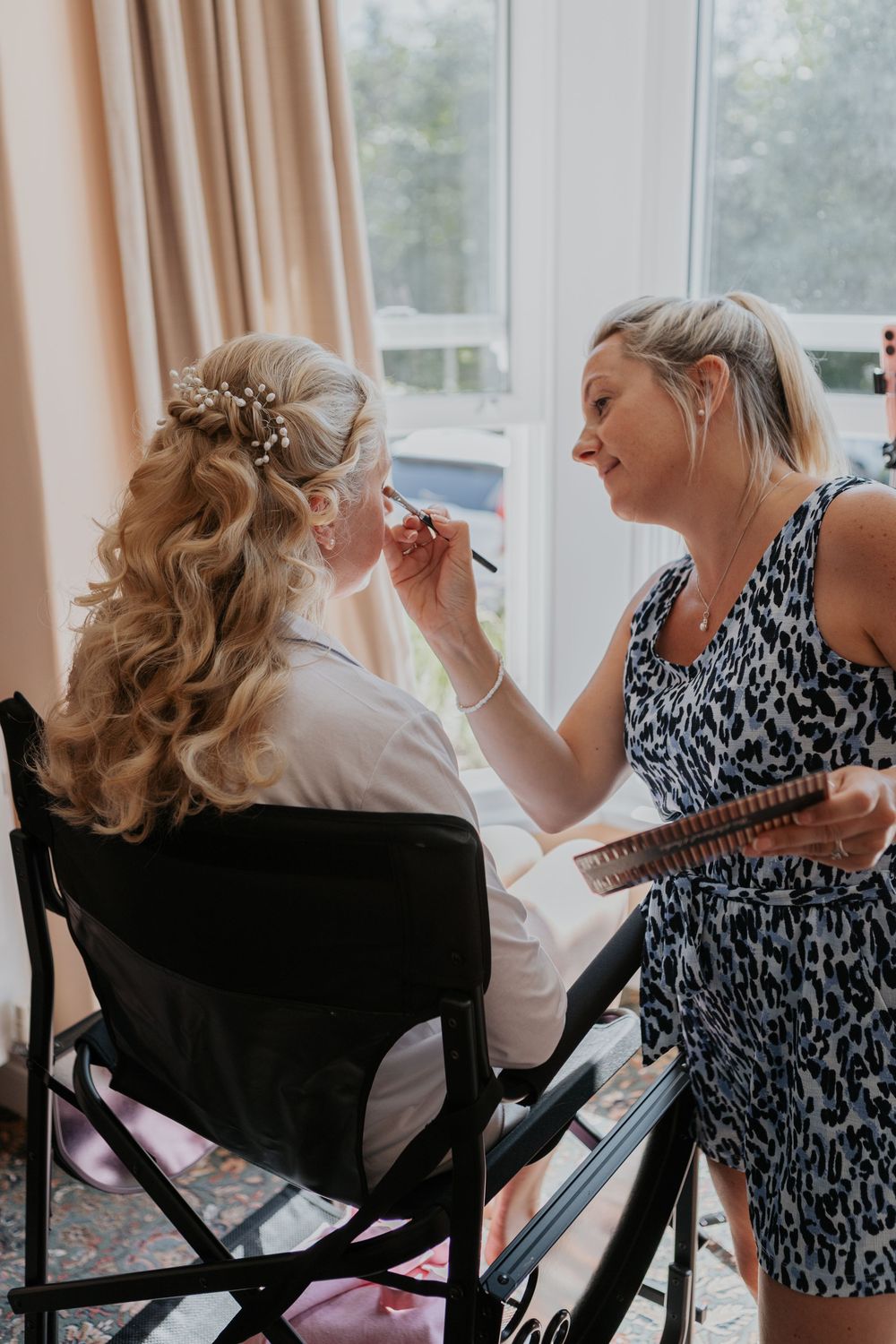 Professional makeup artist applying makeup to a bride with floral hair accessories by a window.