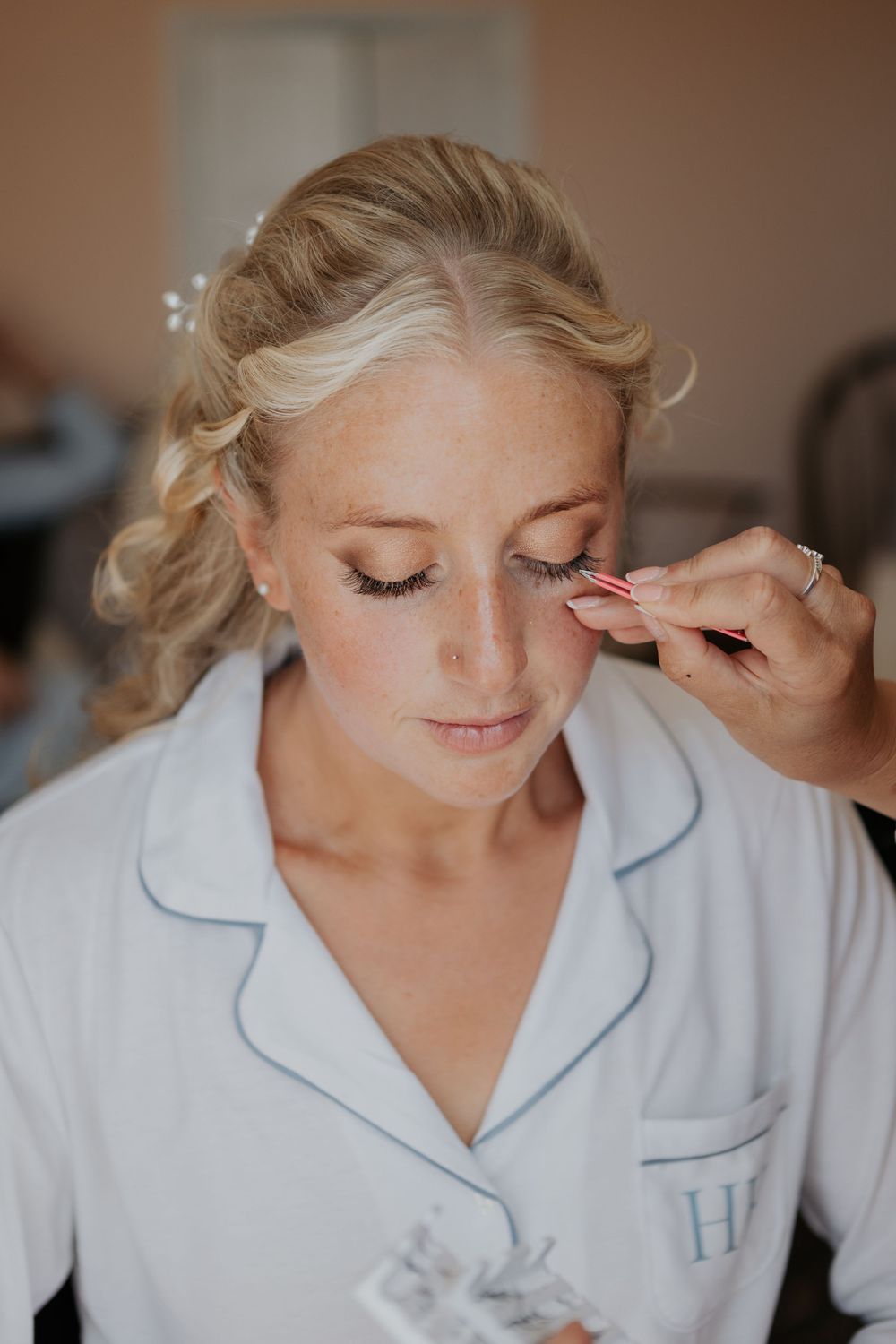 Close-up of a makeup artist applying false lashes to a blonde bride in white monogrammed pajamas.