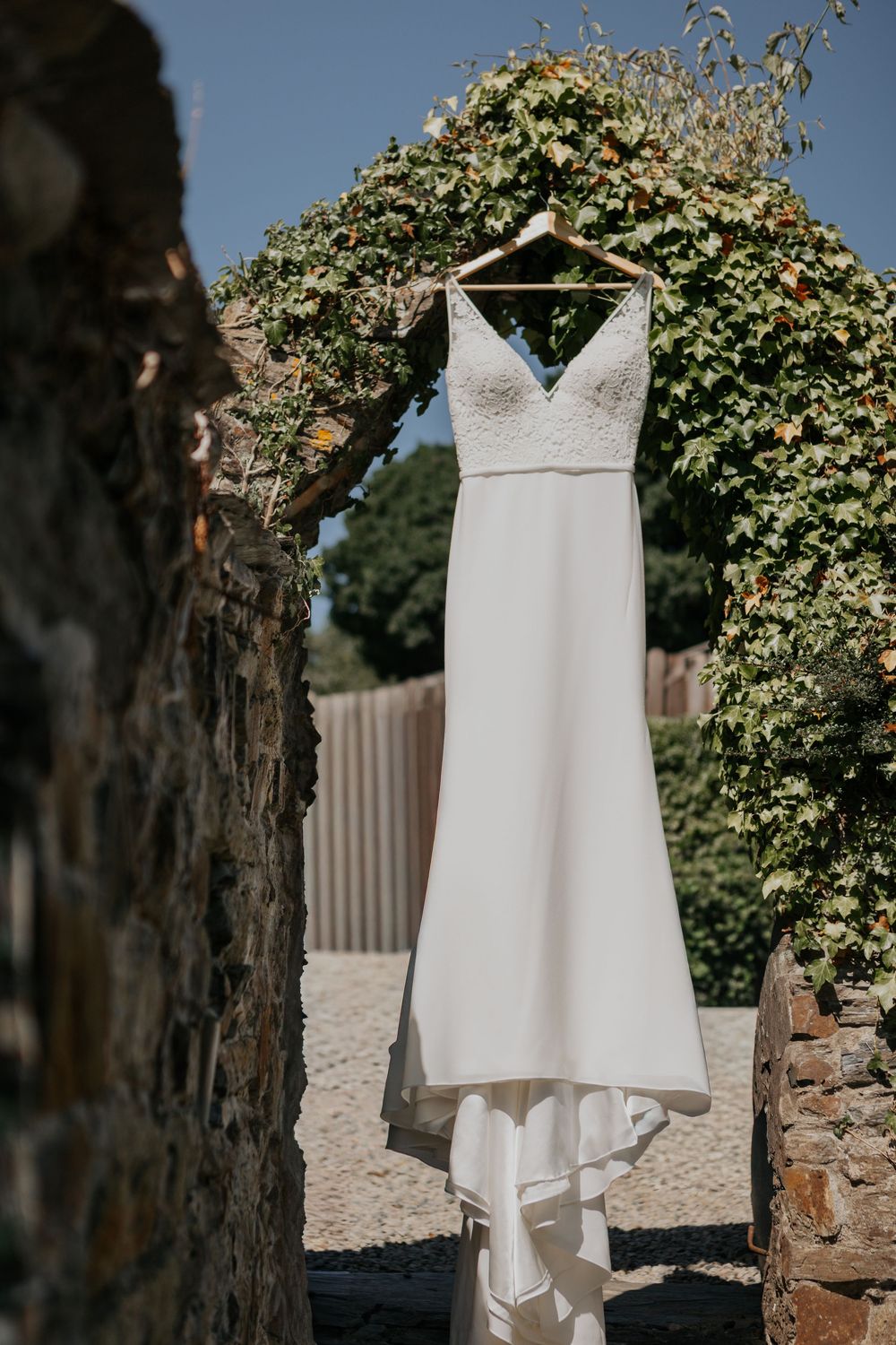Elegant white spaghetti strap wedding dress hanging on a wooden hanger against a rustic stone arch with green ivy.