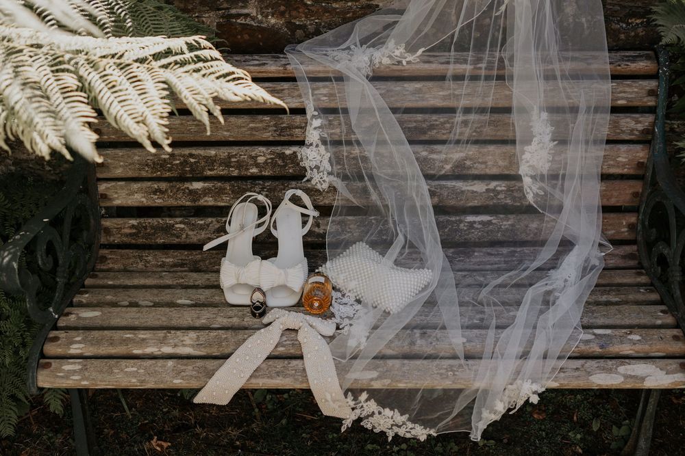 Bridal accessories including white heels, veil, and ribbon bow arranged on a wooden bench with fern leaves.