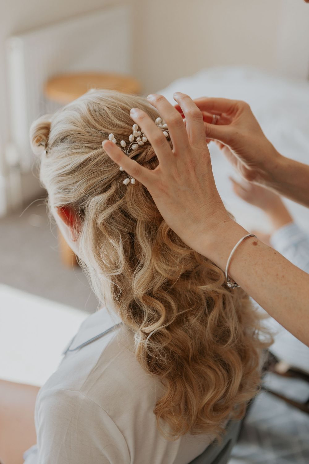 Hands styling a bride's wavy blonde hair with delicate pearl pins during wedding preparations.