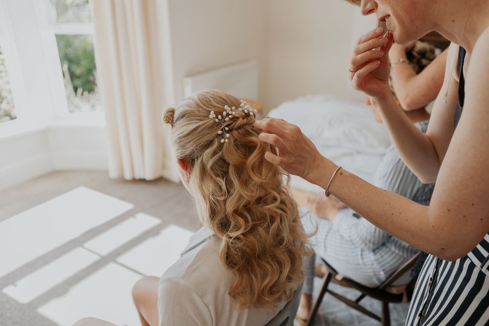 Hairstylist adding pearl hair accessories to a bride's elegant wavy updo in a bright bedroom.