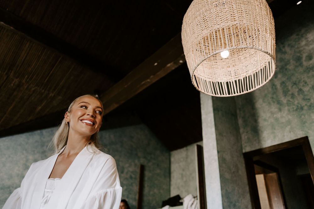 A person in white clothing smiles while standing in a room with decorative hanging light fixtures.