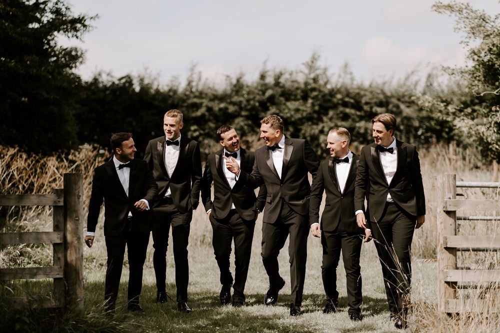 Groomsmen in black tuxedos walking and laughing together outdoors in a natural garden setting.