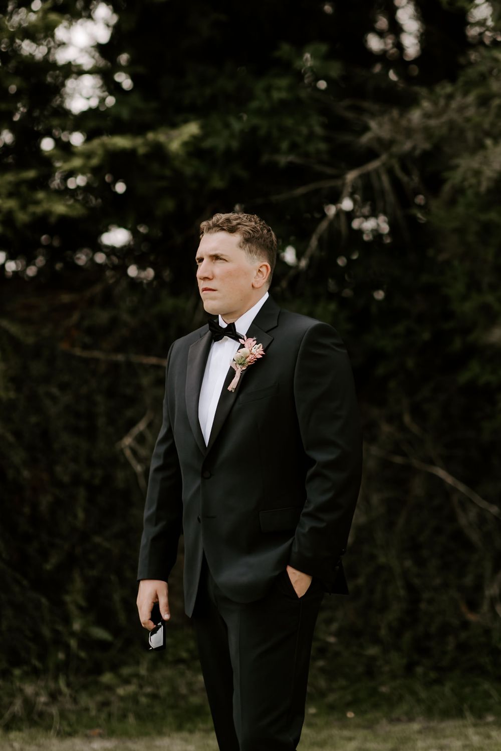 A person wearing a formal black tuxedo stands outdoors against a dark green foliage background.