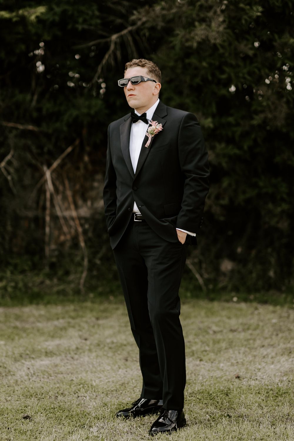 A person in a formal black tuxedo and sunglasses poses outdoors in a natural setting.