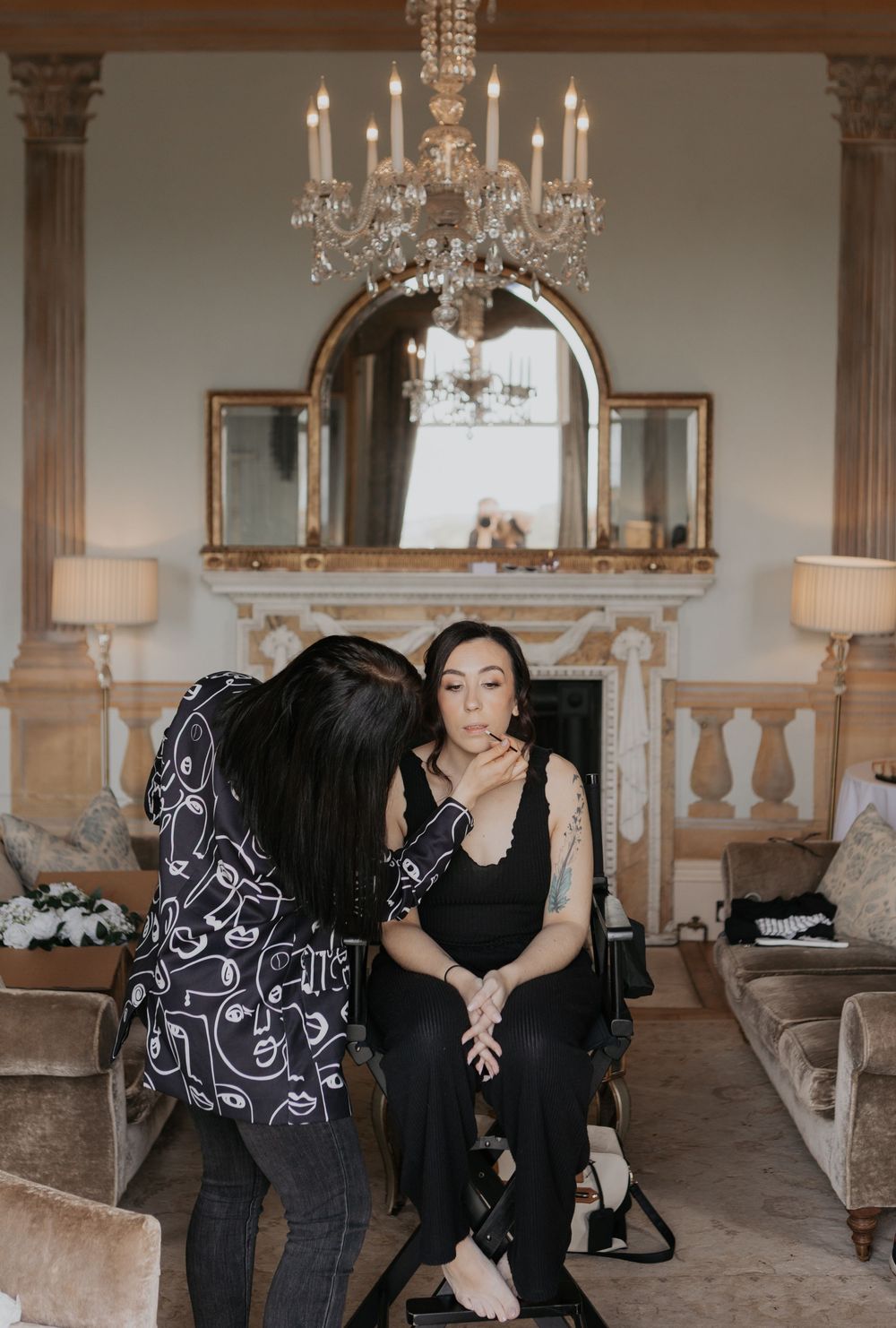 Makeup artist applies finishing touches to bridesmaid in black dress under crystal chandelier in ornate room.