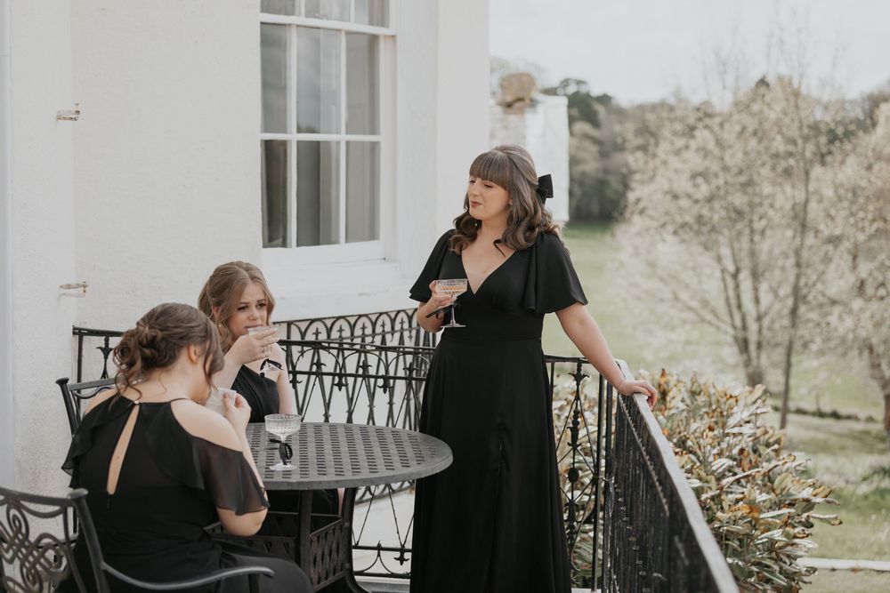 Three bridesmaids in black dresses relax on ornate balcony with scenic garden views on wedding day.