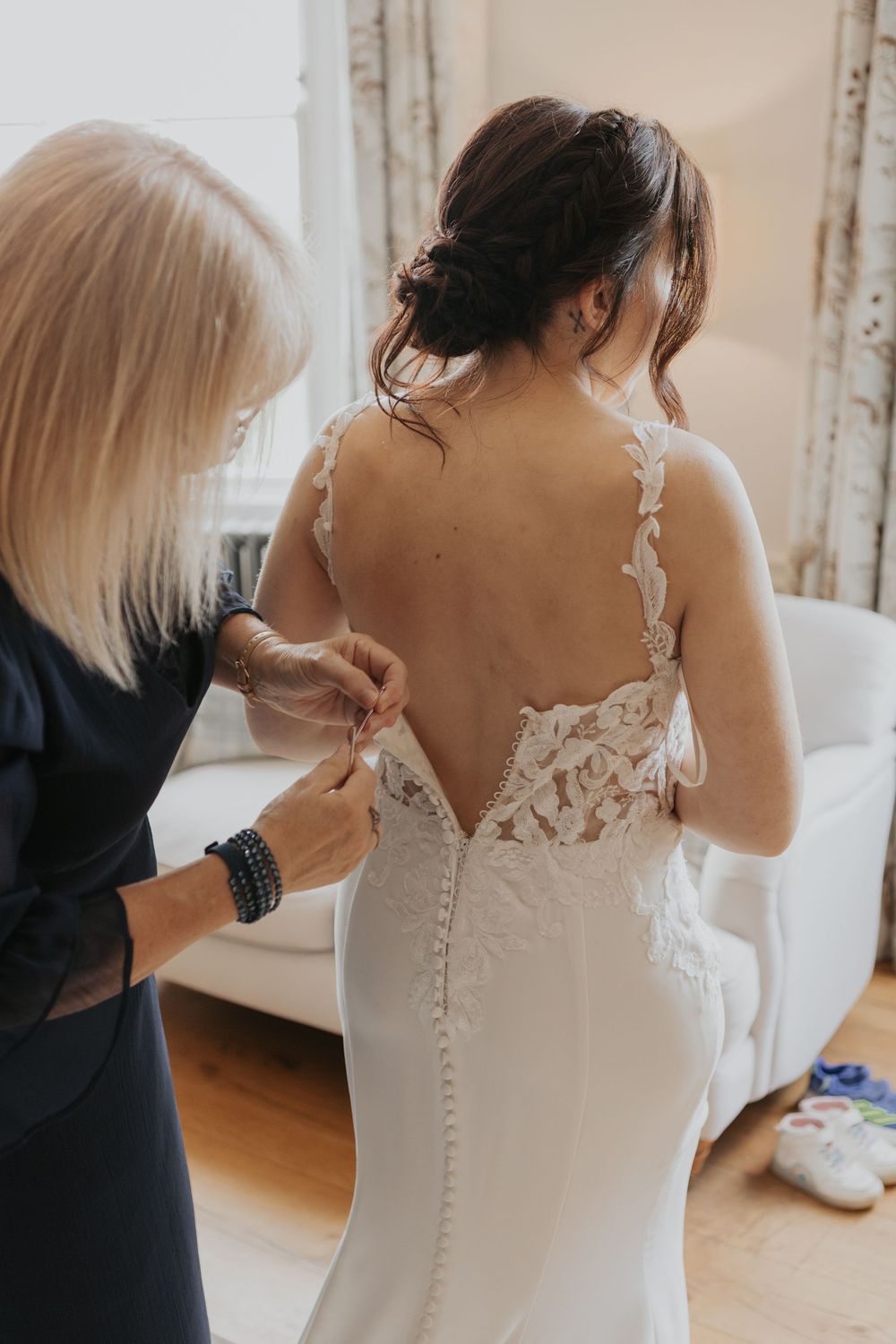 Mother fastens delicate lace buttons on brides stunning backless wedding gown with floral lace detail.