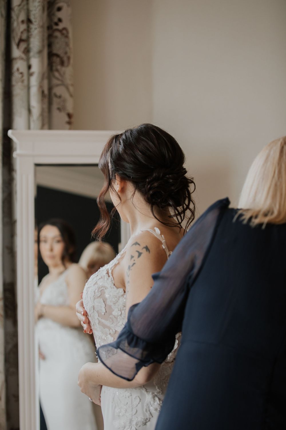 Bride with updo and shoulder tattoo views her lace wedding gown in full-length mirror with assistance.
