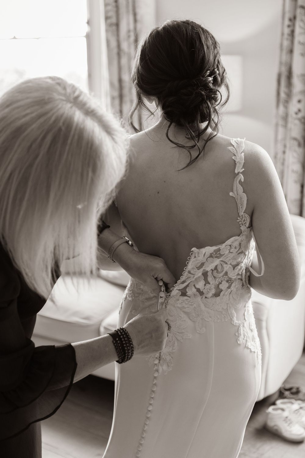 Black and white image of mother carefully buttoning brides elegant lace backless wedding dress.