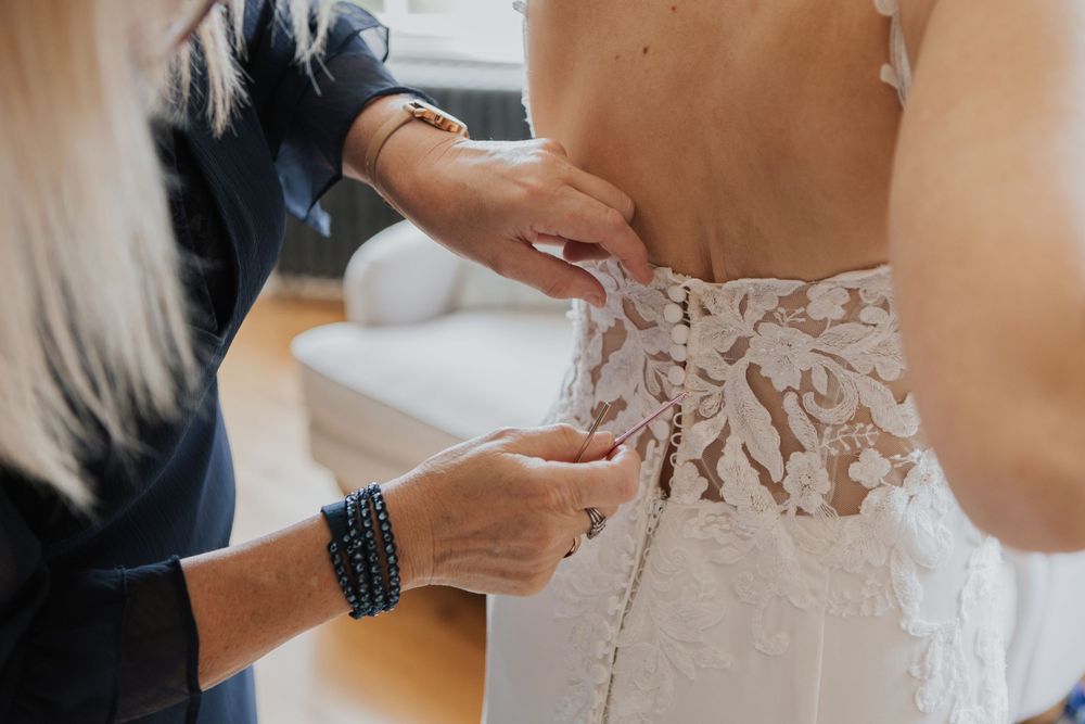 Helper fastening intricate lace buttons on back of bridal gown during wedding preparation.