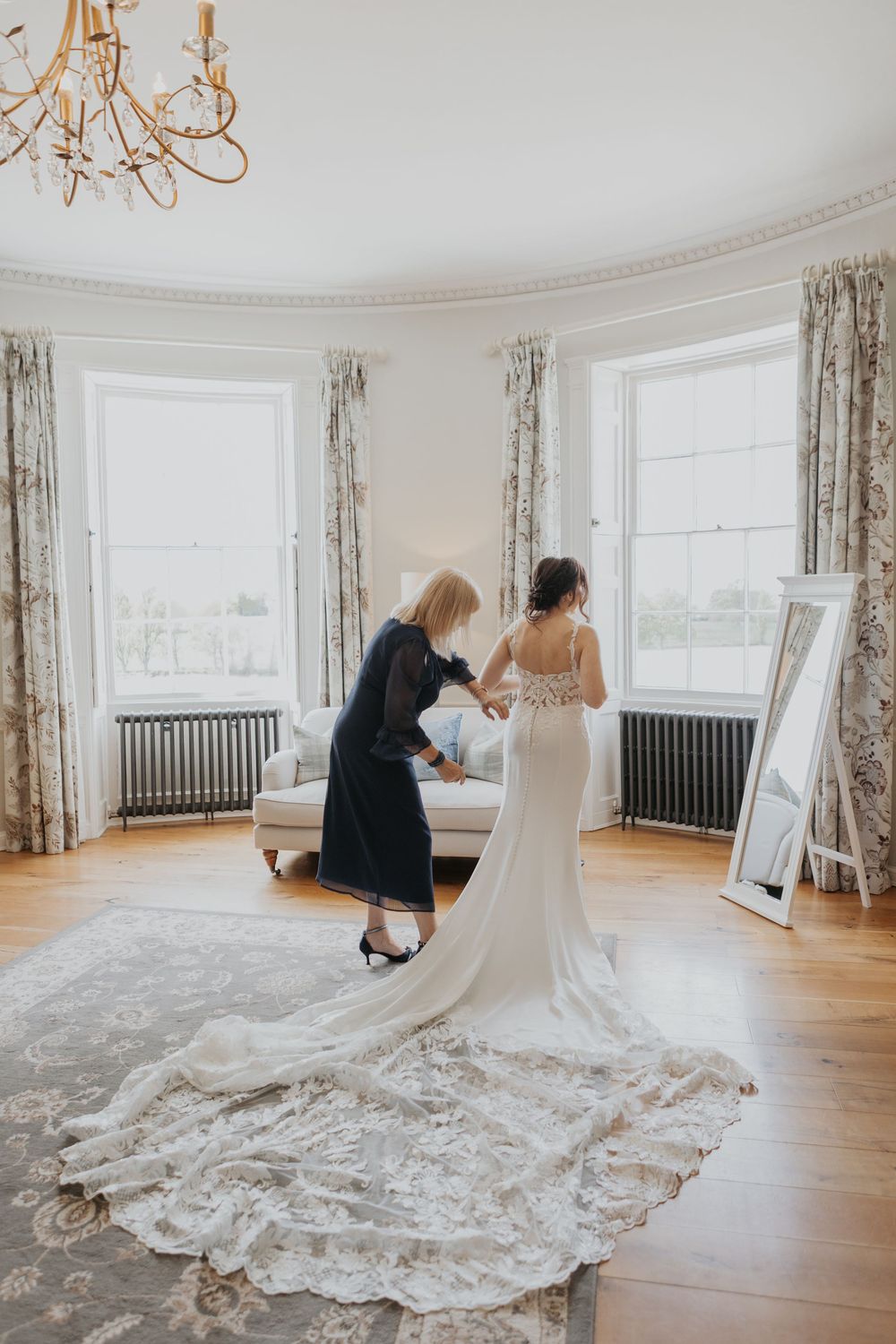 Bride in flowing lace train gown stands in bright bay window room as mother fastens her dress.