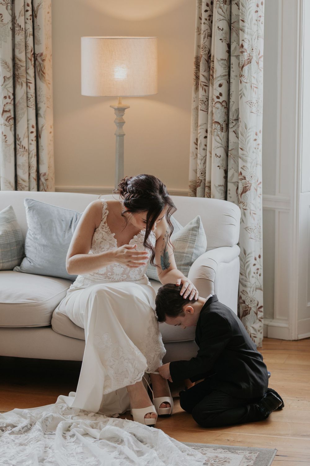 Groom kneeling to help bride put on wedding shoes while she sits on elegant white sofa.