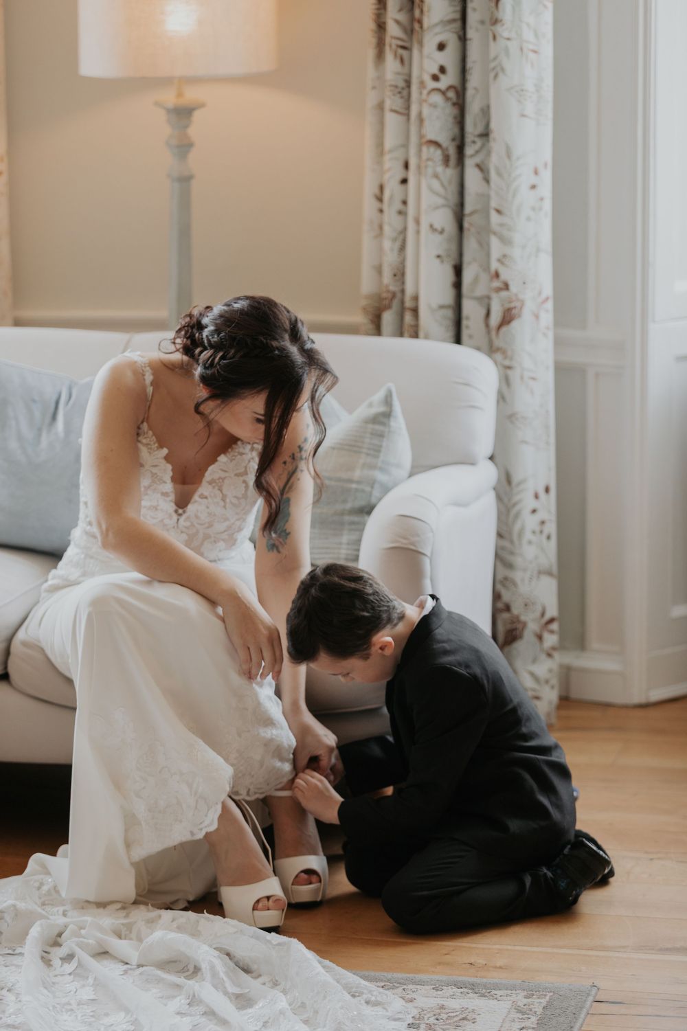 Groom carefully fastening bride's white heels as she sits on sofa in lace wedding dress.