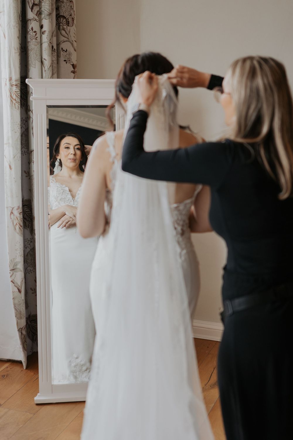 Wedding assistant placing veil on bride's hair as she views reflection in ornate floor mirror.