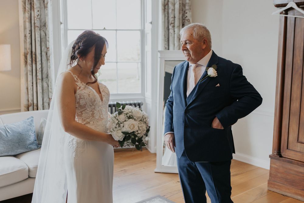 Bride in lace wedding gown sharing first look moment with father in elegant room.