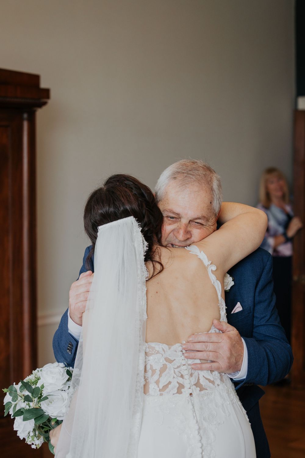 Emotional father embracing bride in lace backless wedding gown before wedding ceremony.