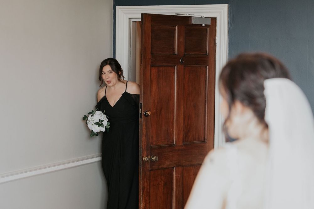 Bridesmaid in black dress holding white bouquet watching bride during wedding preparations.