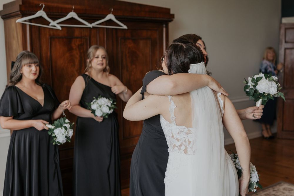 Bridesmaids in black dresses reacting emotionally as they see bride in lace wedding gown.