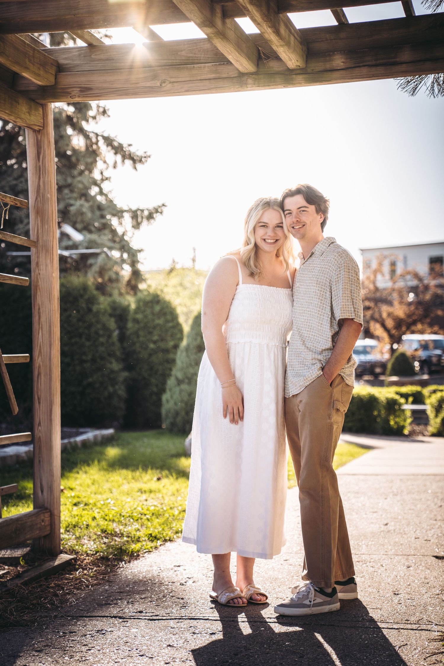 A smiling couple poses under a wooden pergola in a sunny garden, woman in white dress, man in casual shirt and khakis.