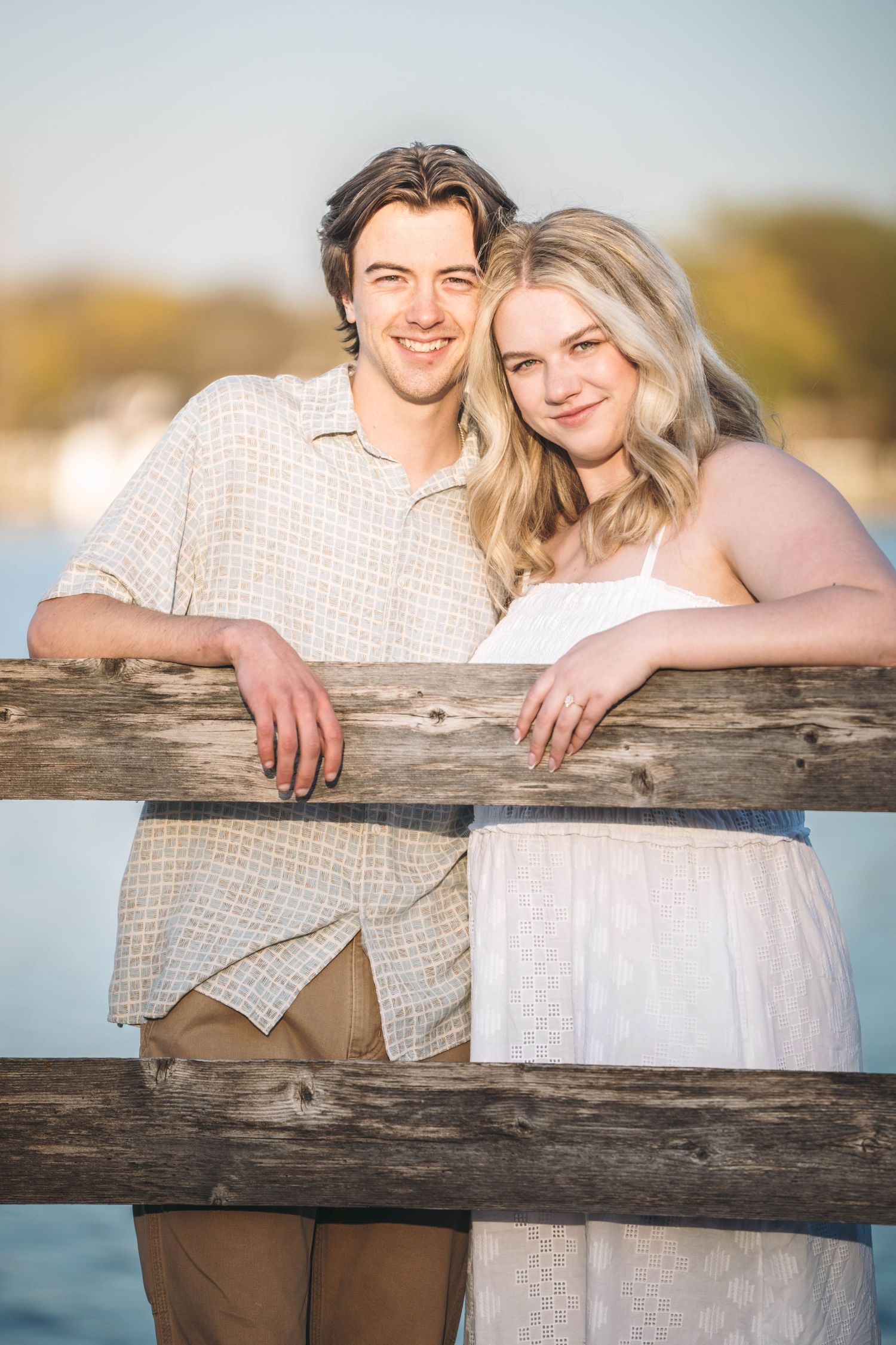 Smiling young couple leaning on a wooden fence by the water during golden hour.