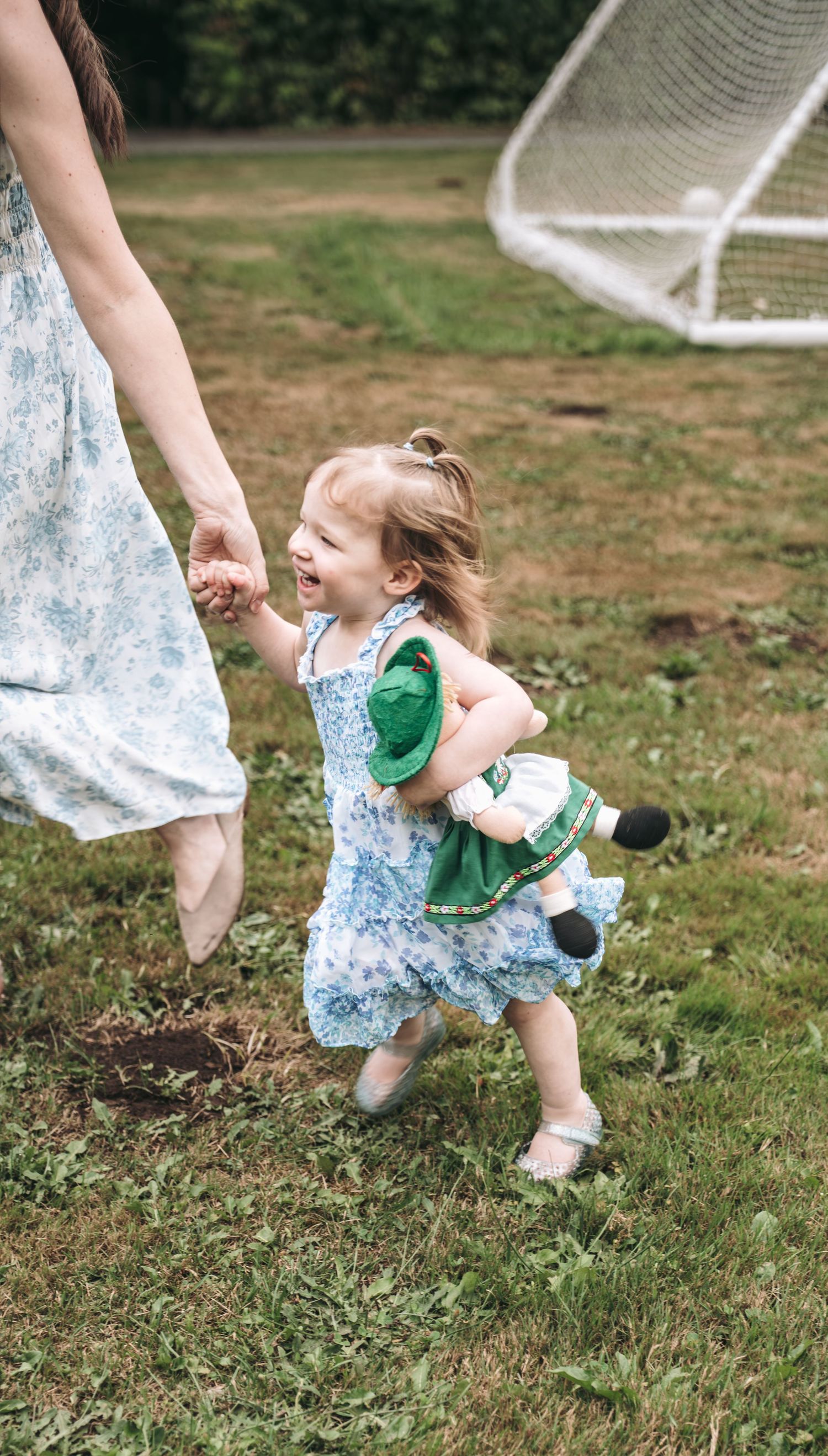 A joyful toddler girl in a floral dress holds her mother's hand while running outdoors, clutching a green stuffed toy.