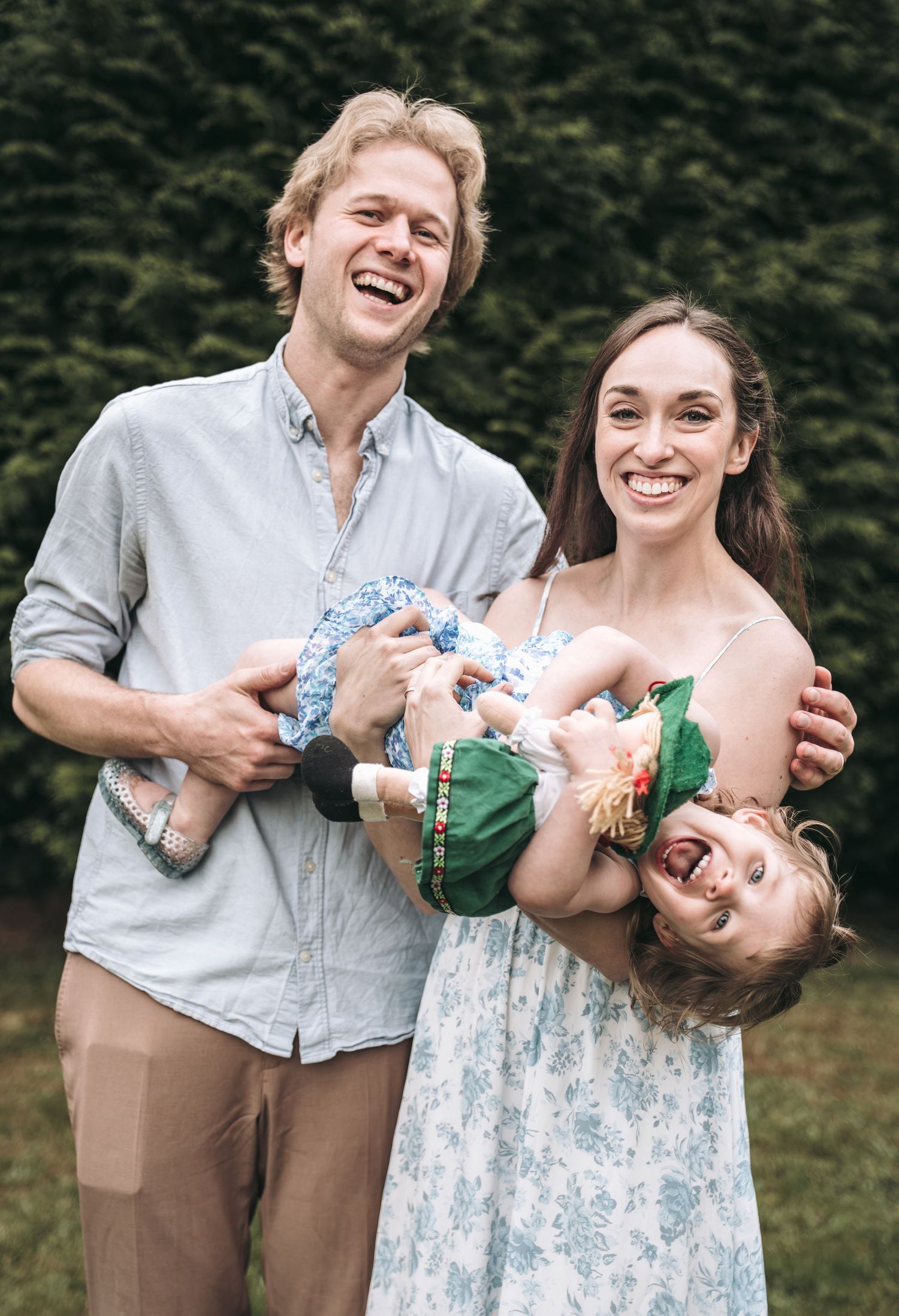 Happy family with laughing parents holding a playful toddler upside down outdoors near green hedges.