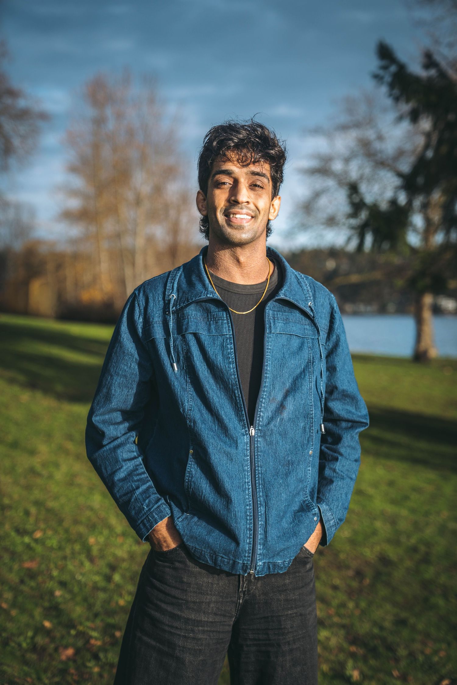 Smiling young man in a denim jacket standing confidently in a sunny green park.