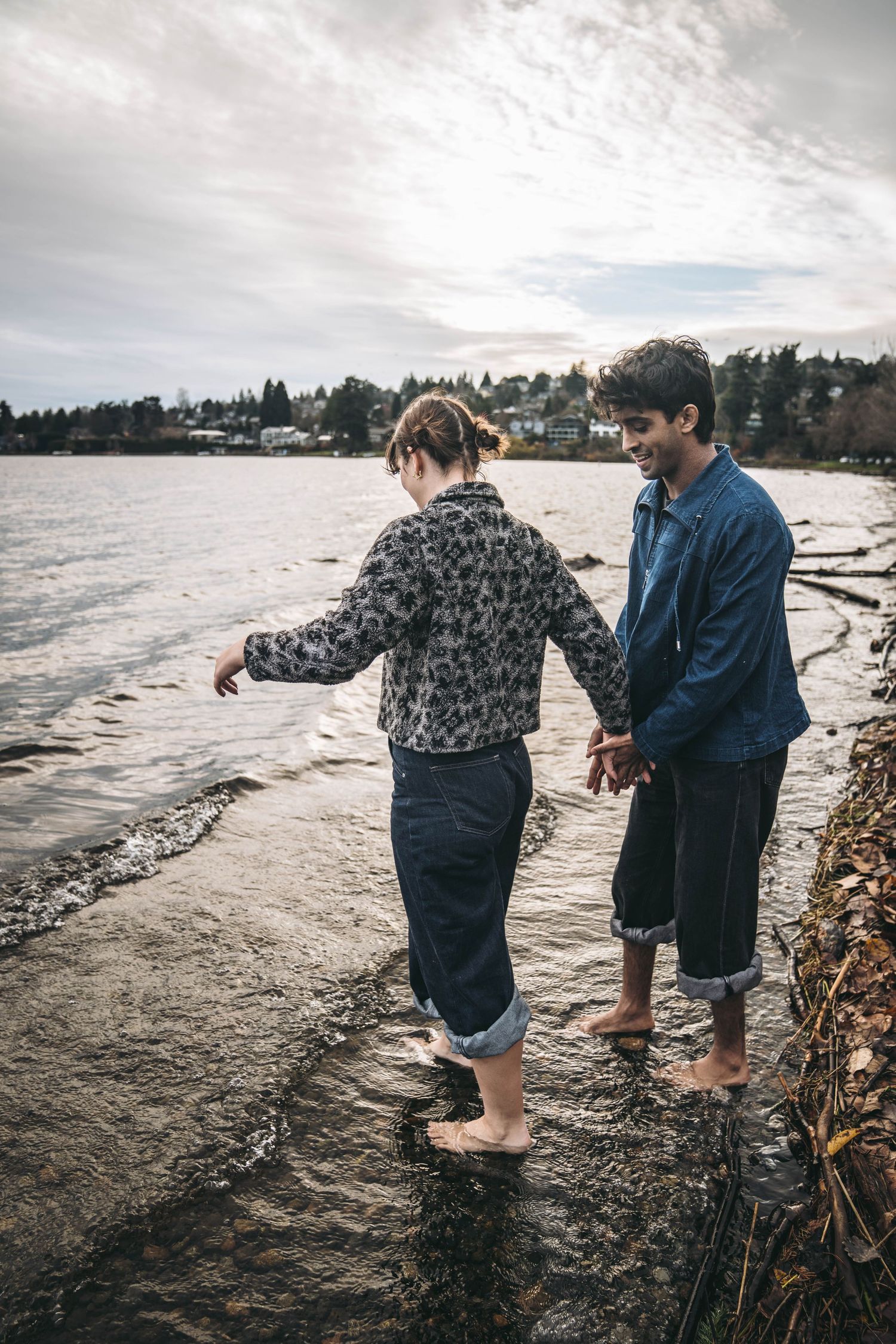 Couple holding hands and wading barefoot along a scenic rocky shoreline at sunset.