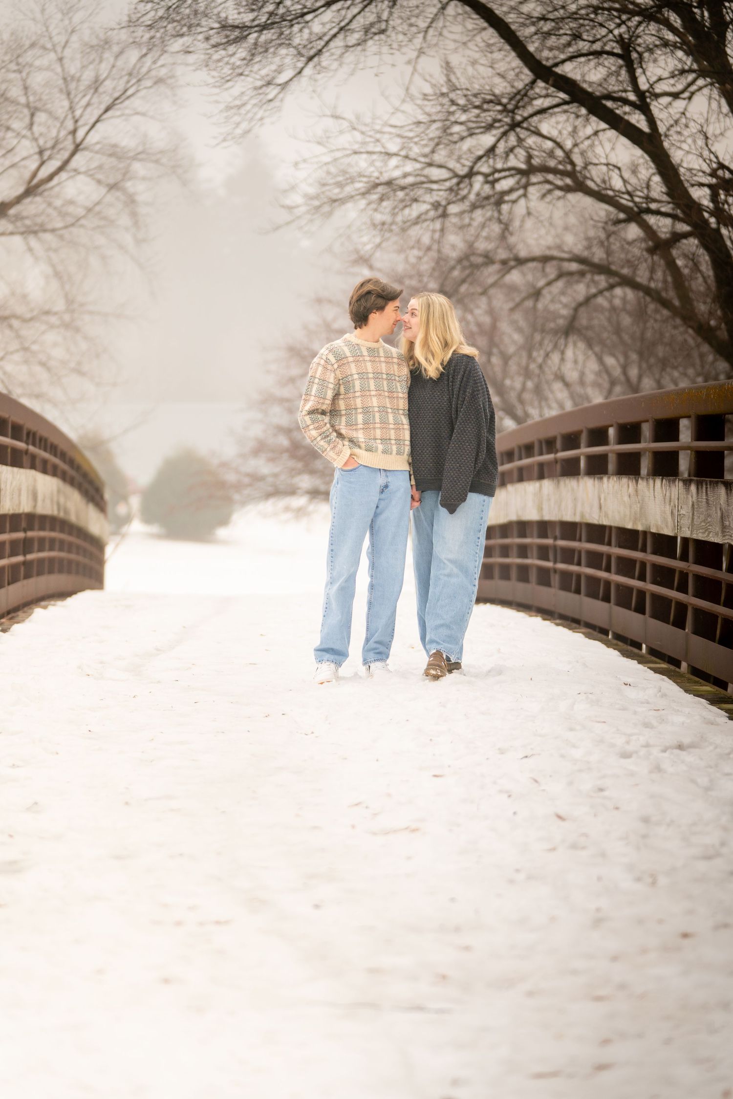 Couple walking hand in hand on a snowy bridge surrounded by bare winter trees.