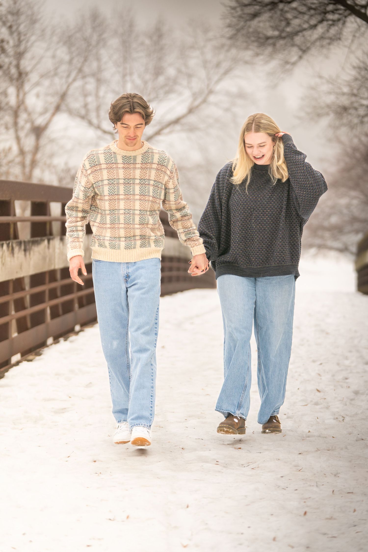 Young couple holding hands and laughing while walking on a snow-covered bridge in winter.