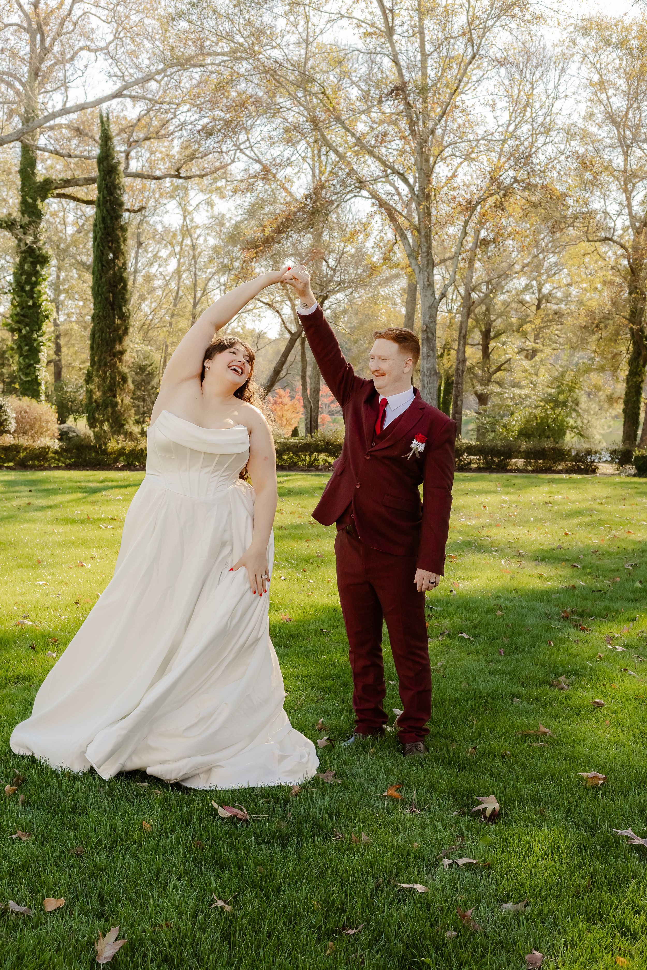 A couple in wedding attire dances together on a grassy lawn surrounded by autumn trees in a romantic outdoor setting.