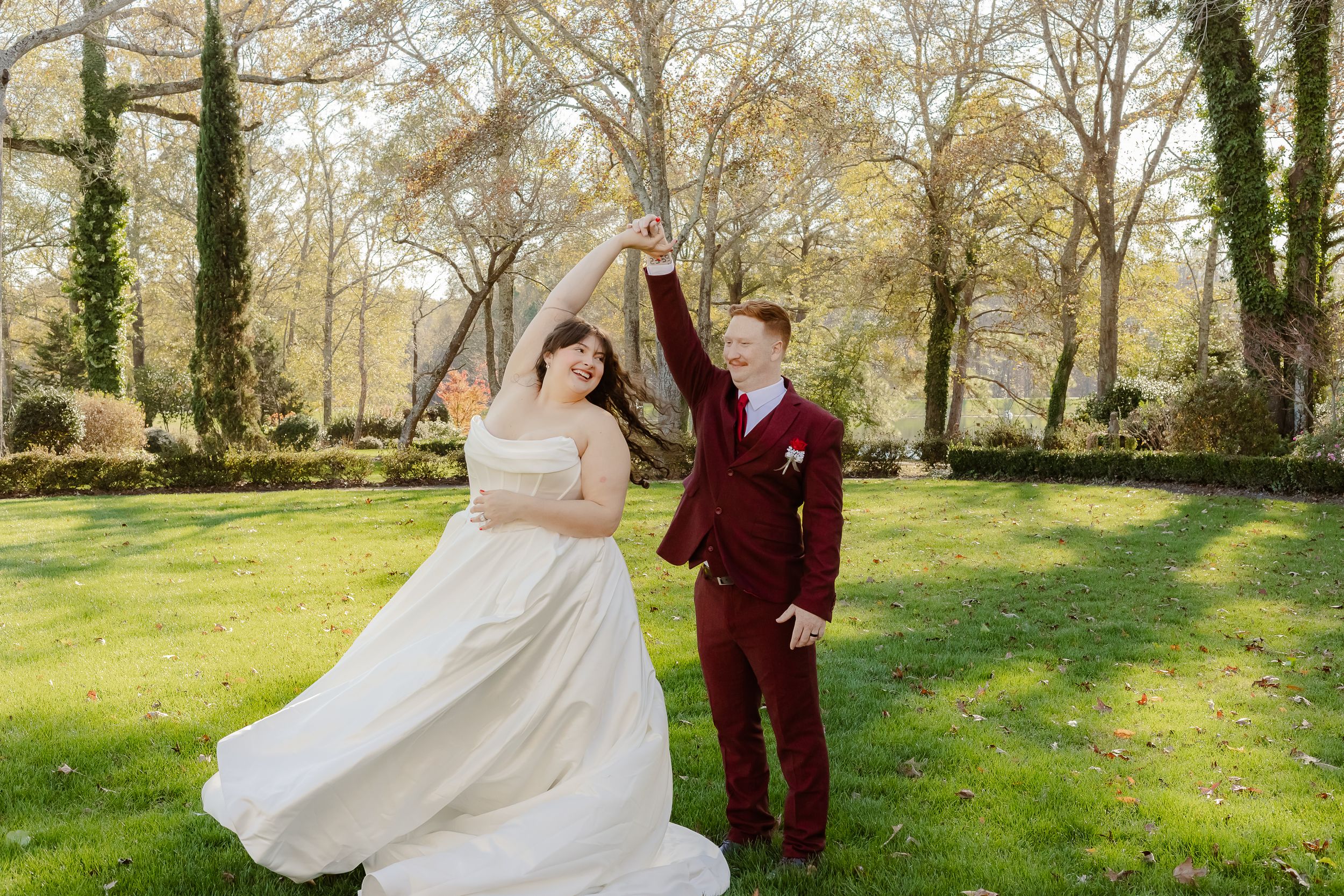 A couple in wedding attire dances and twirls in a sunlit park with spring trees in the background.