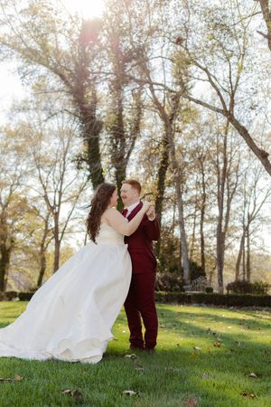 A couple in wedding attire embrace in a sunlit autumn forest setting with golden light filtering through the trees.