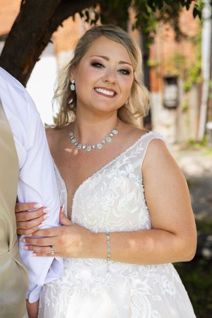 Elegant bride in white lace dress with crystal jewelry poses against brick wall.