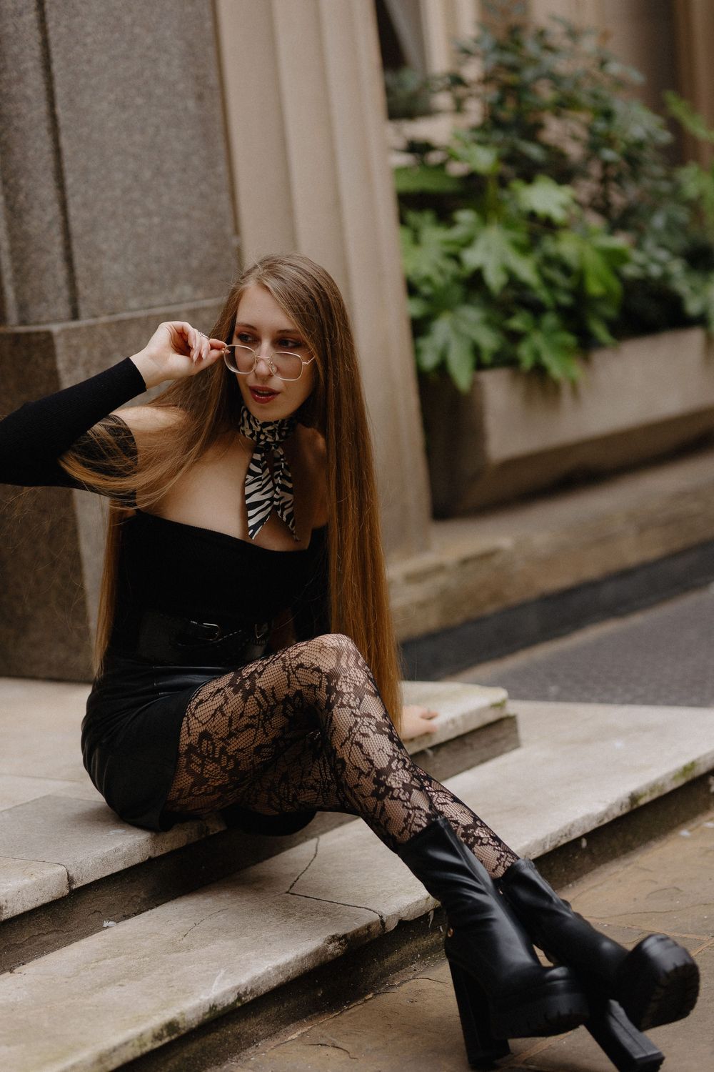 Fashion portrait of someone in black lace dress and boots sitting on stone steps in an outdoor setting.