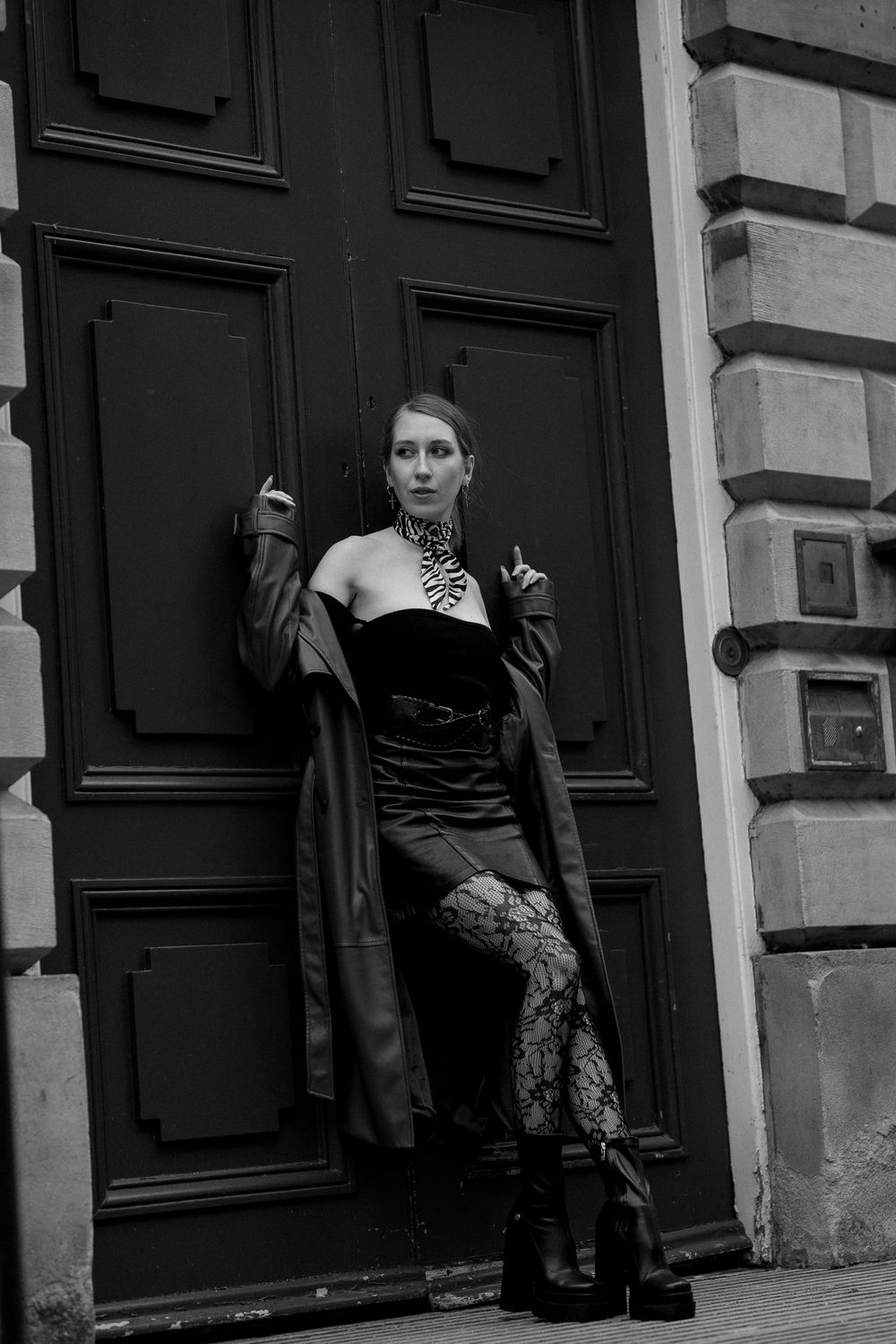 Woman in dark clothing poses against black doors in moody black and white photography.