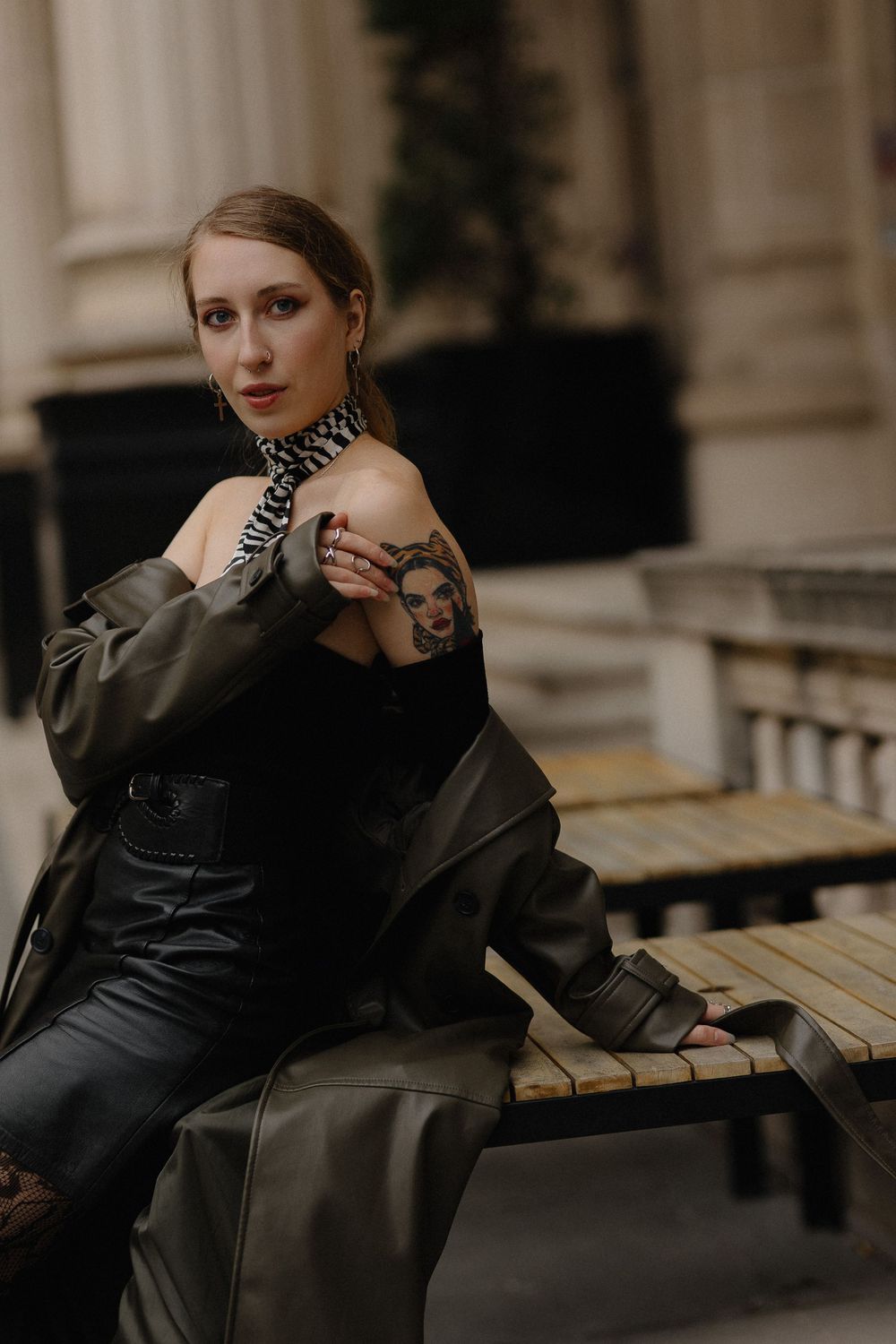 A figure in a black coat sits thoughtfully on a wooden bench against a classical architectural backdrop.