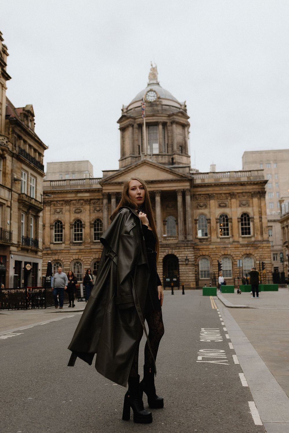 A figure in a black coat poses in front of a grand historic building with classical architecture and domed features.