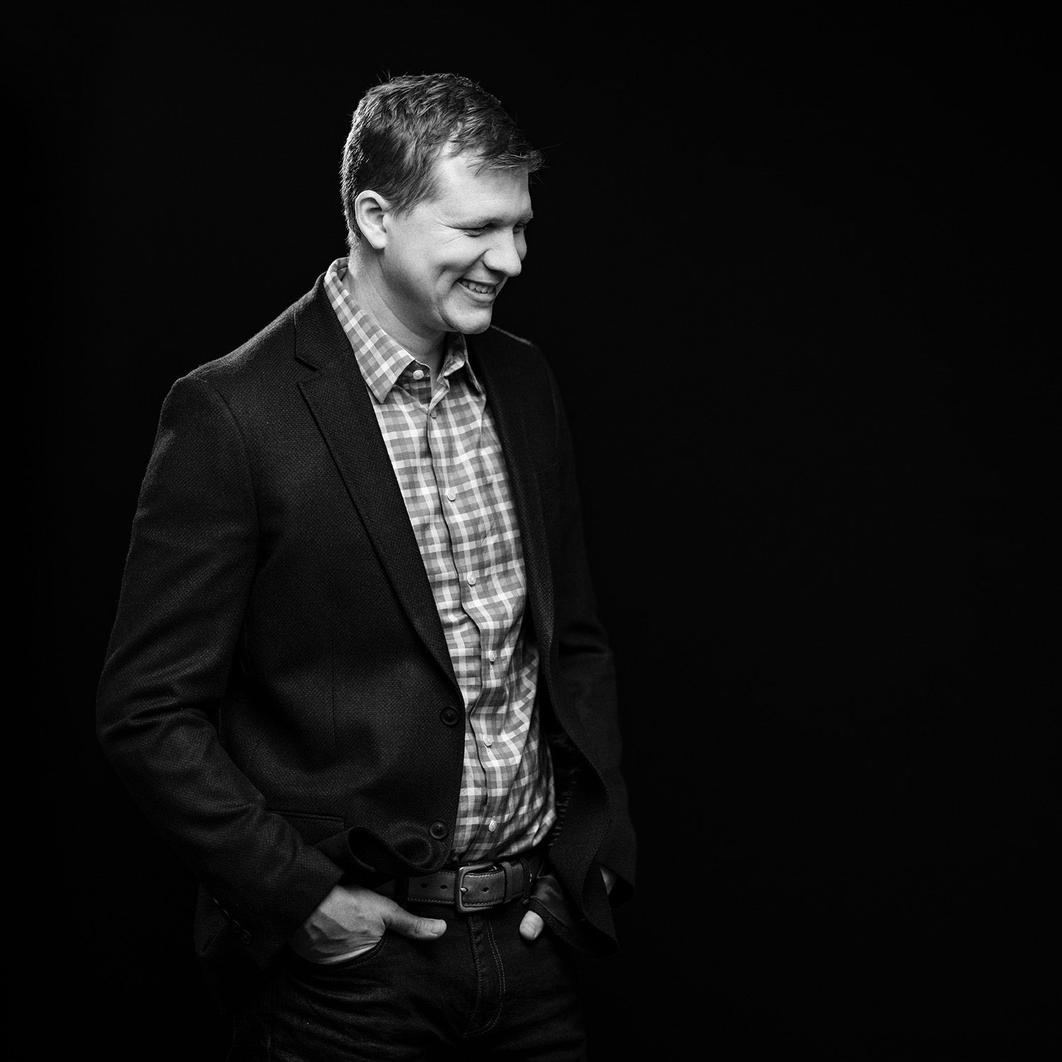 Person in dark suit jacket and checkered shirt stands against black background in dramatic lighting.
