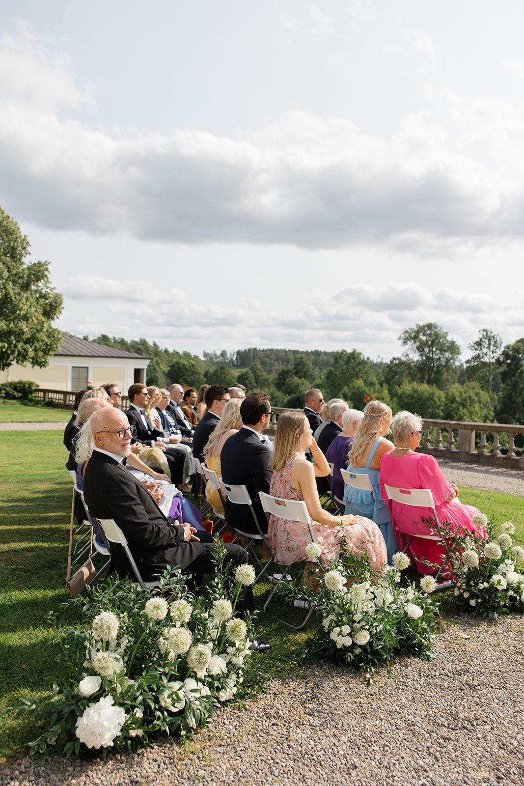Wedding guests seated in rows on gravel path with white floral arrangements lining the aisle during outdoor ceremony.