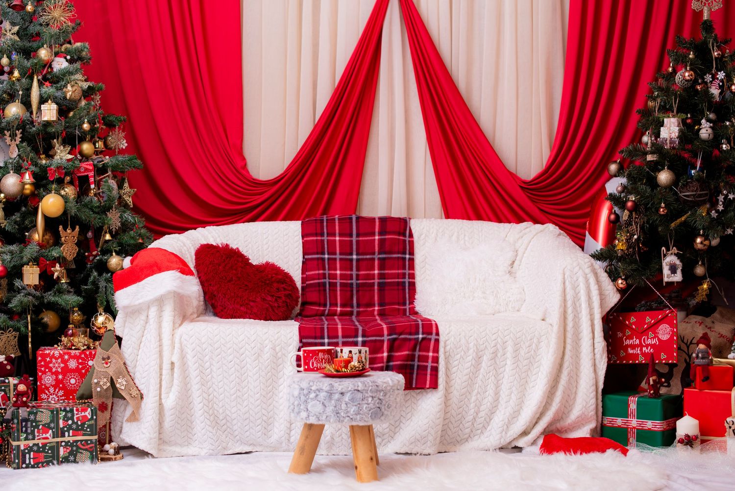 Christmas photography setup with red drapes, white couch, plaid blanket, and decorated trees in the background.